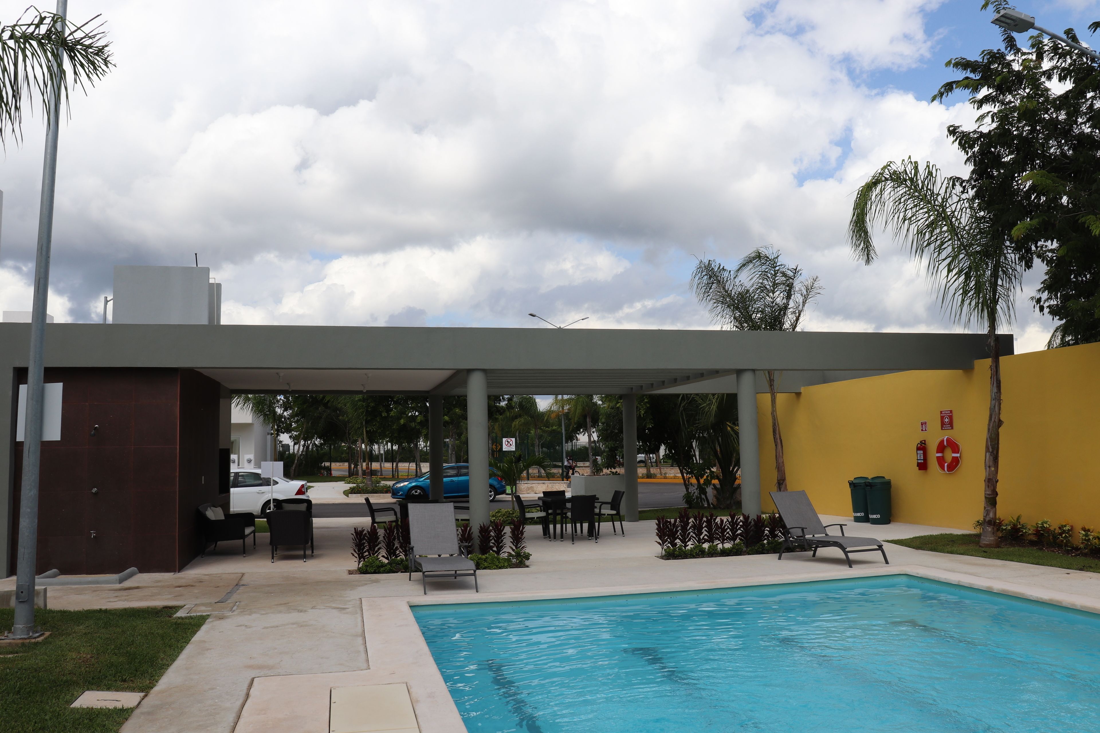 Piscina con sillones de descanso y un área de estar cubierta, rodeada de palmeras y un muro amarillo.