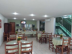 A restaurant with round tables and wooden chairs, a kitchen area, a television, and a staircase visible through a glass wall.