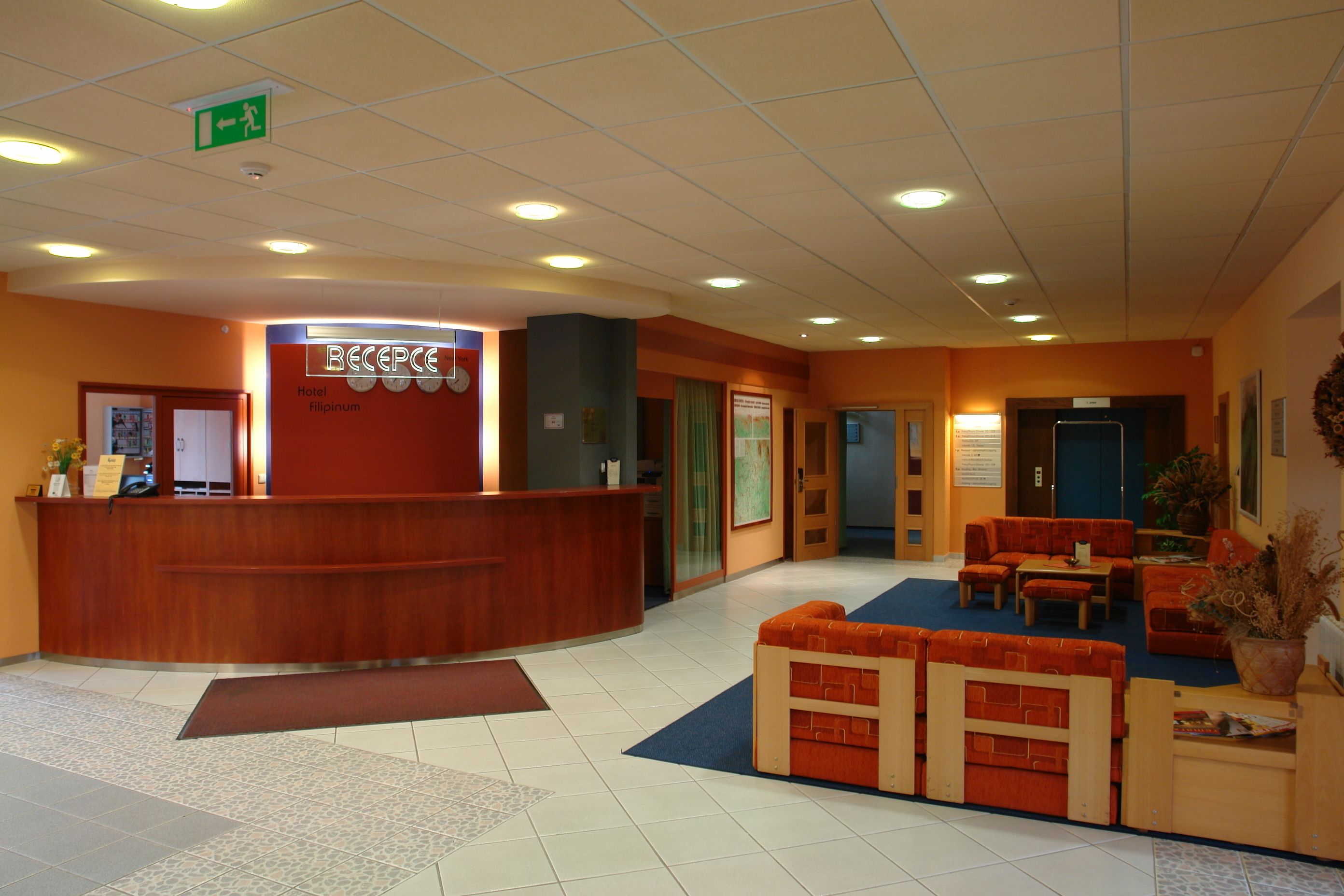 A hotel reception area with a wooden reception desk, a seating area with orange sofas, and a blue carpet.