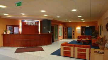 A hotel reception area with a wooden reception desk, a seating area with orange sofas, and a blue carpet.