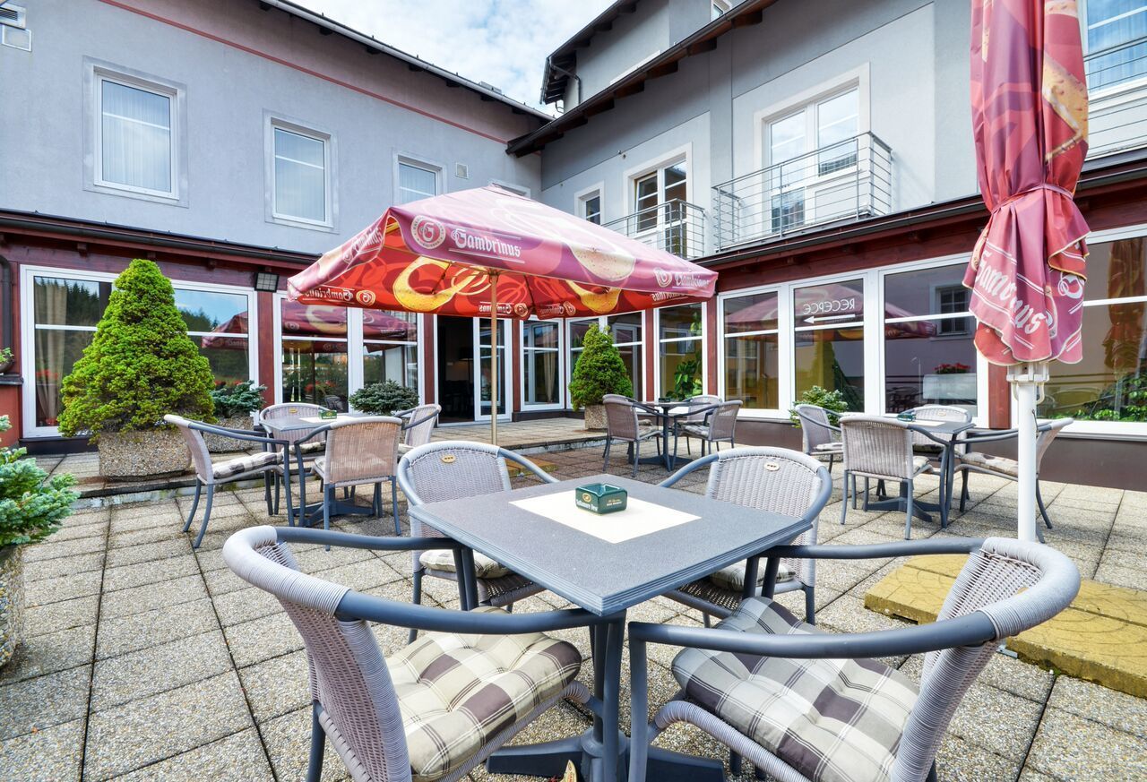 An outdoor patio with wicker furniture and a table, surrounded by a building with large windows and a red umbrella.