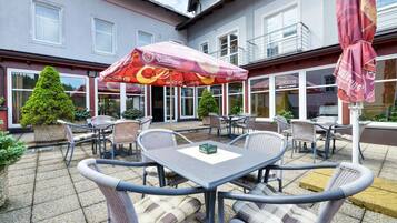 An outdoor patio with wicker furniture and a table, surrounded by a building with large windows and a red umbrella.