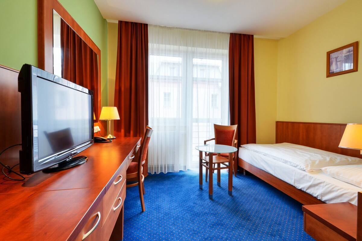 A hotel room with a bed, a desk with a television, a chair, and a window with curtains.