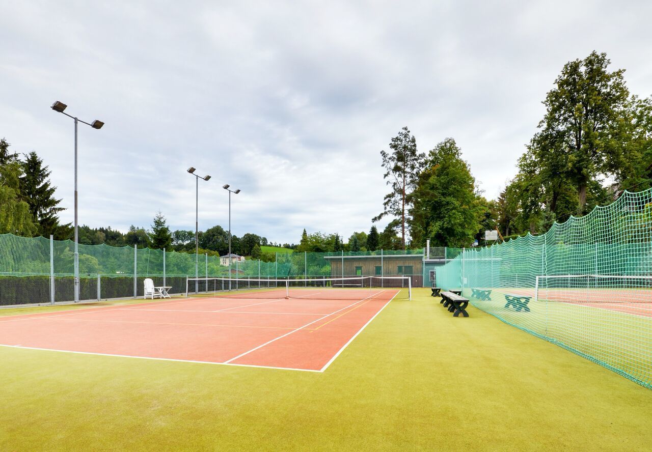 A tennis court with a net, floodlights, and seating areas.