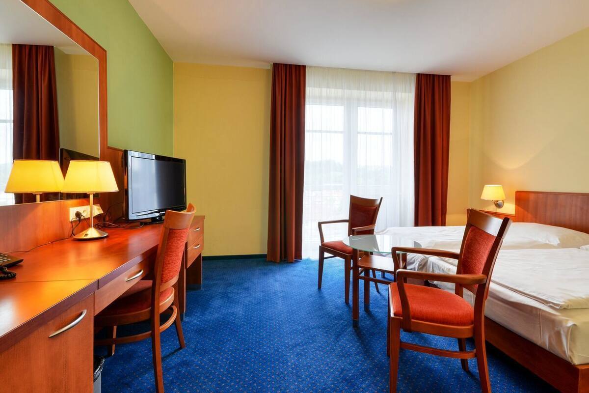 A hotel room with a desk, two chairs, a bed, a television, and a window with curtains.