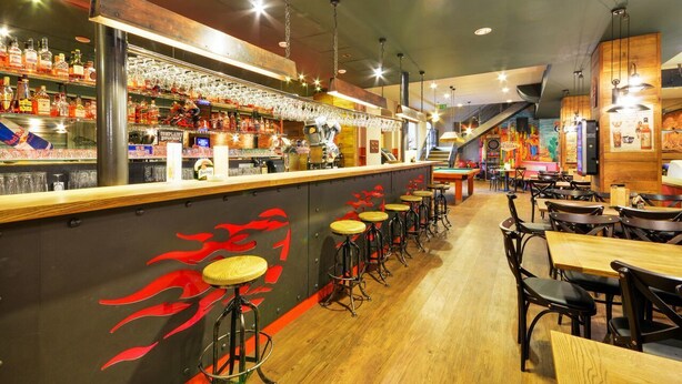 A bar area with stools, a counter with bottles, and a mural with red flames. - Hotel Filipinum (Jablonne nad Orlici)