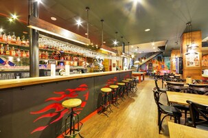 Una zona de bar con taburetes, una barra repleta de botellas y un mural con llamas rojas.