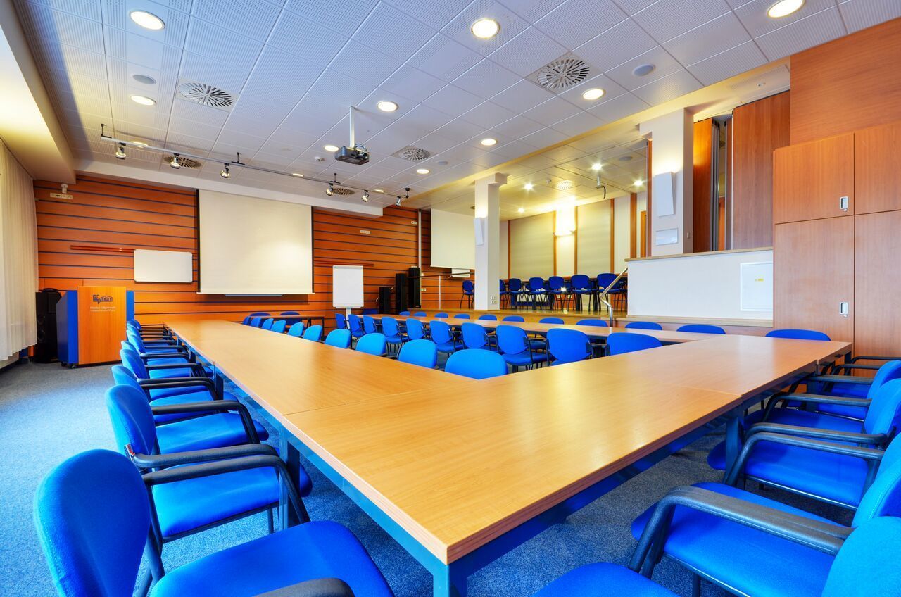 Ein Konferenzraum mit einem langen Holztisch, umgeben von blauen Stühlen, einer Whiteboard und einer Projektionsleinwand.