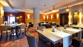 Une salle à manger d’hôtel avec plusieurs tables et chaises, un sol recouvert de moquette et des luminaires suspendus.