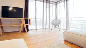 A modern hotel room with a flat-screen TV on a wooden stand, a round chair, a sofa, and sheer curtains.