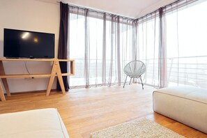 A modern hotel room with a flat-screen TV on a wooden stand, a round chair, a sofa, and sheer curtains.