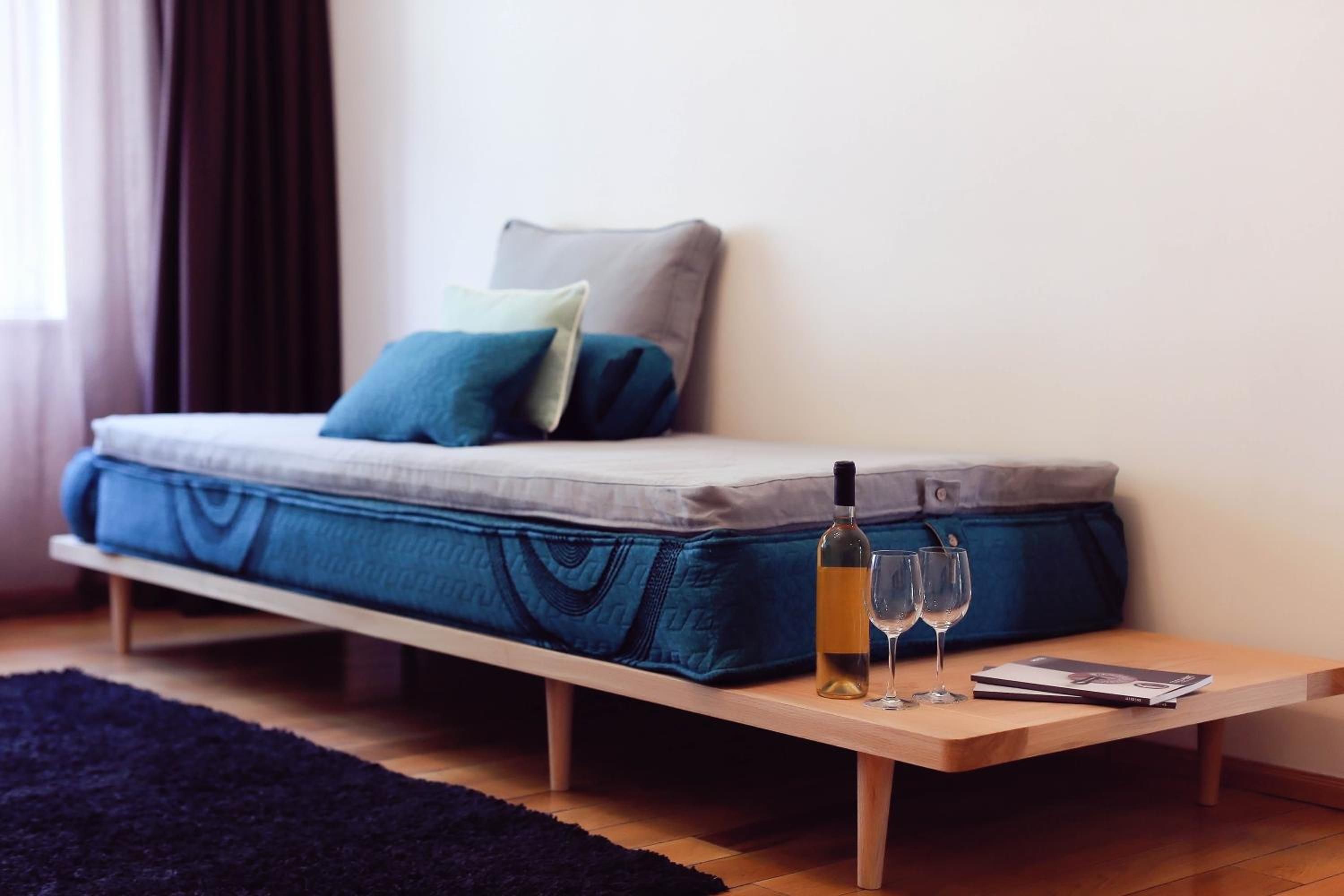 A bed with a mattress, pillows, a bottle of wine, two glasses, and a book on a wooden table.