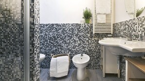 Un baño con pared de mosaicos, un lavamanos blanco, un bidé y una cesta de mimbre con una toalla.