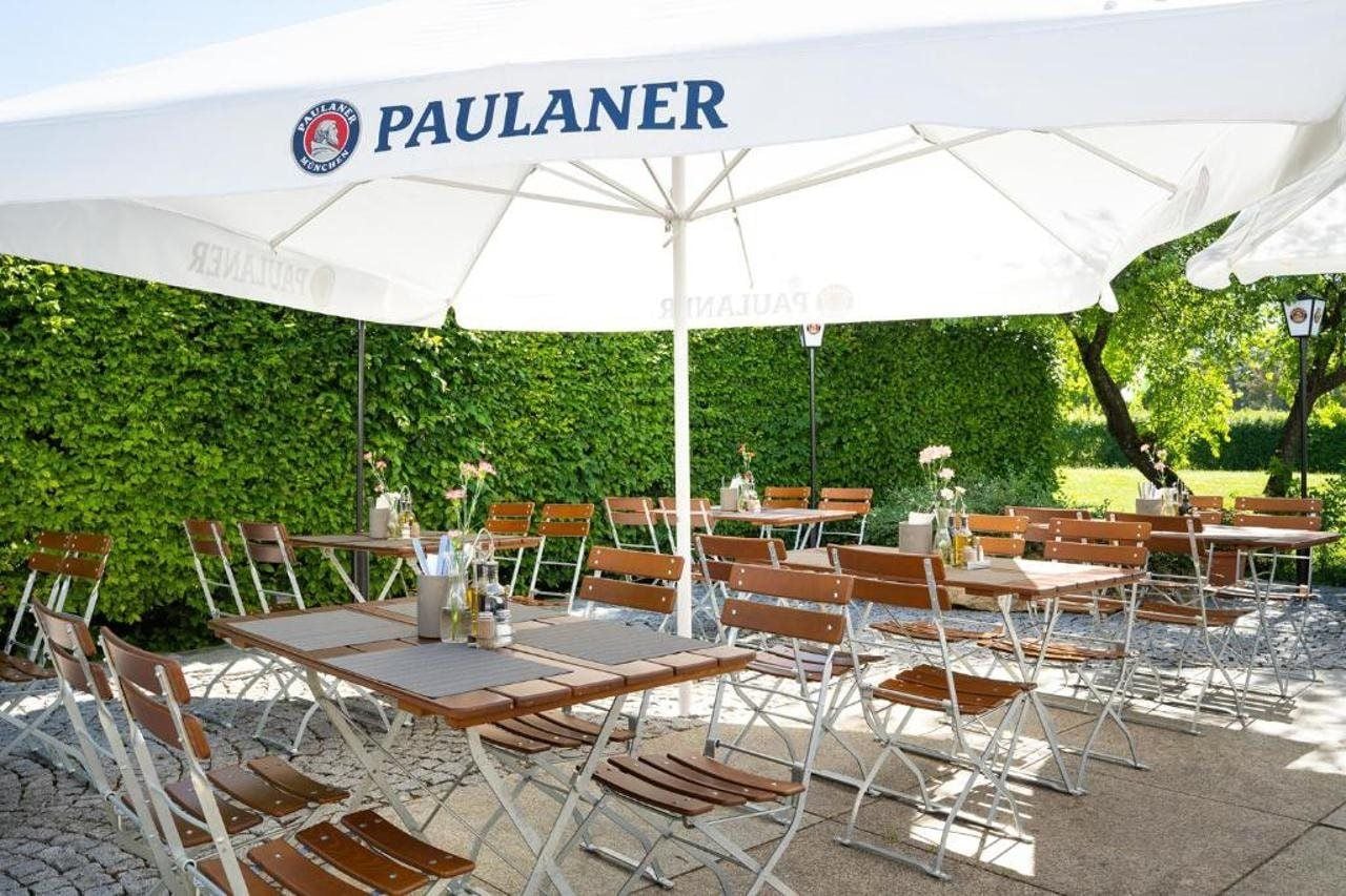 Zona de estar al aire libre con mesas y sillas bajo un gran paraguas blanco con la marca 'PAULANER' a la vista.