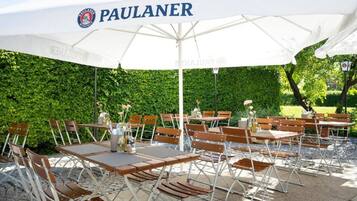 Zona de estar al aire libre con mesas y sillas bajo un gran paraguas blanco con la marca 'PAULANER' a la vista.