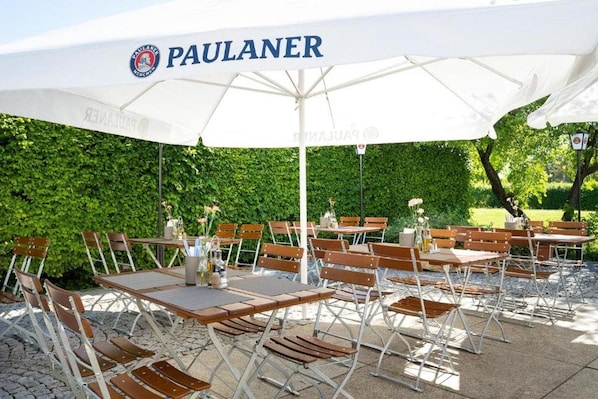 Zona de estar al aire libre con mesas y sillas bajo un gran paraguas blanco con la marca 'PAULANER' a la vista.