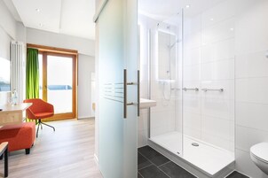 A modern hotel room with a glass shower enclosure, a wooden floor, a red chair, and a view through a large window.