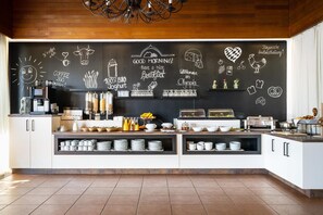 Una cocina moderna con un muro de pizarra que muestra productos para el desayuno y mensajes, una cafetera y vajilla de desayuno ordenada.