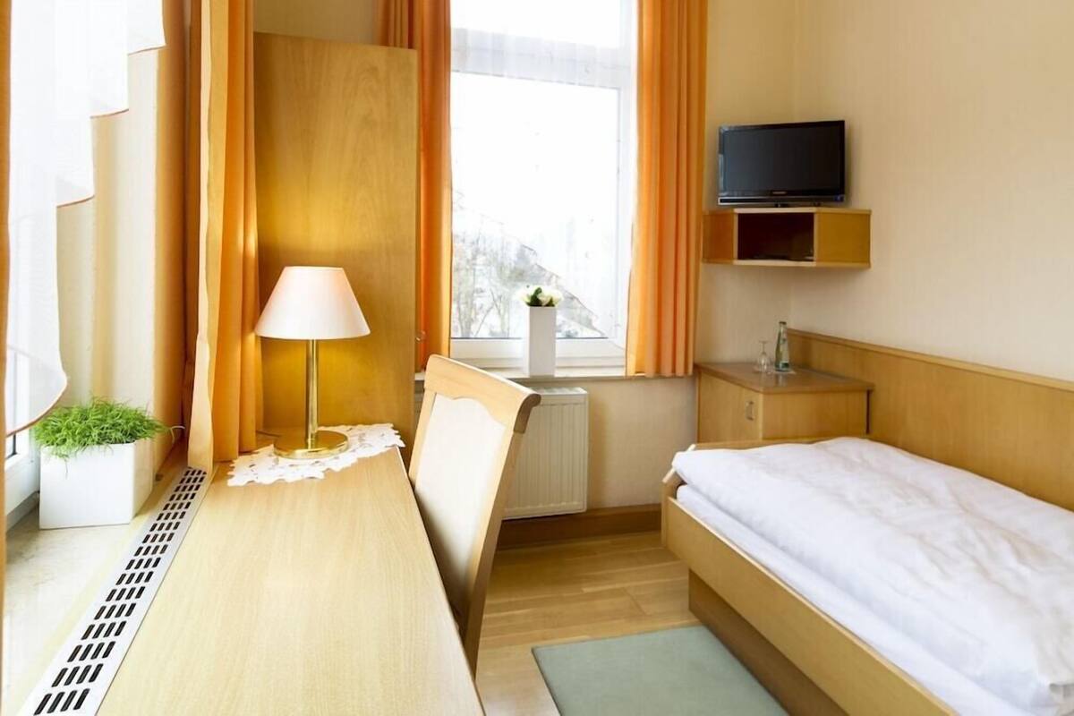 A hotel room with a bed, a desk with a lamp, a television on a shelf, a radiator, and a window with curtains.