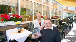Ein Straènkencafé mit Tischen und Stühhlen, großen Fenstern mit Spitzenvorhängen und Blumentopfpflanzen.