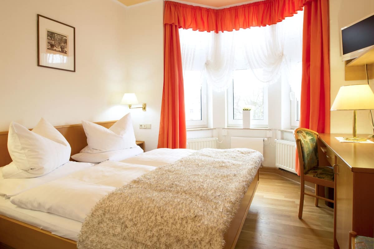 A hotel room with a bed, a desk, and a chair. There is a window with curtains and a picture on the wall.