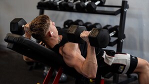 Una persona ejercitándose en un banco con mancuernas, en un gimnasio con una barra de pesas al fondo.