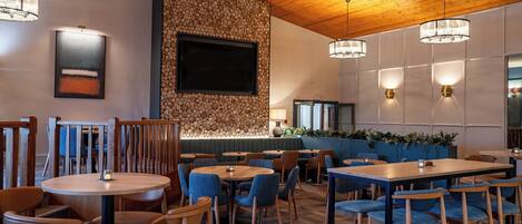 Un restaurant avec des tables rondes et des chaises bleues, un plafond en bois et un mur de pierre avec un téléviseur.