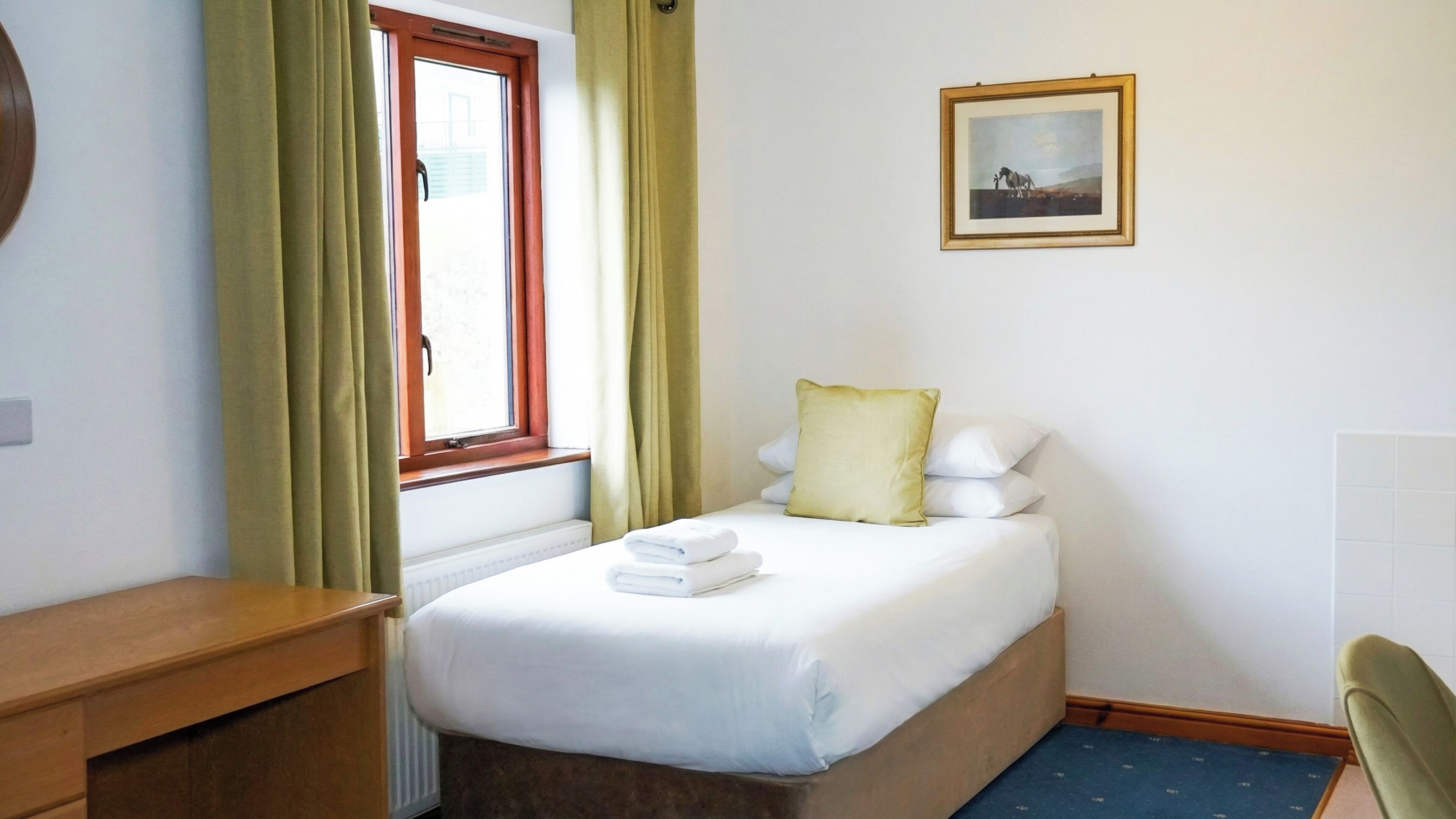 Una habitación de hotel pequeña y ordenada, con una cama individual, una mesita de noche de madera, una silla y un cuadro en la pared.