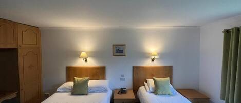 Une chambre d’hôtel avec deux lits simples, chacun doté d’une table de chevet et d’une lampe. Il y a une armoire en bois sur la gauche et une fenêtre avec des rideaux sur la droite.
