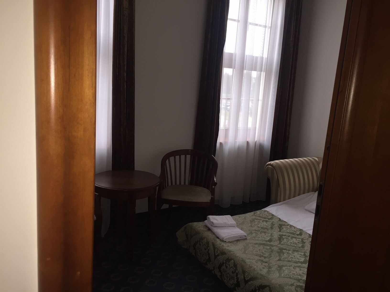 A hotel room with a bed, a chair, a small table, and a window with curtains.
