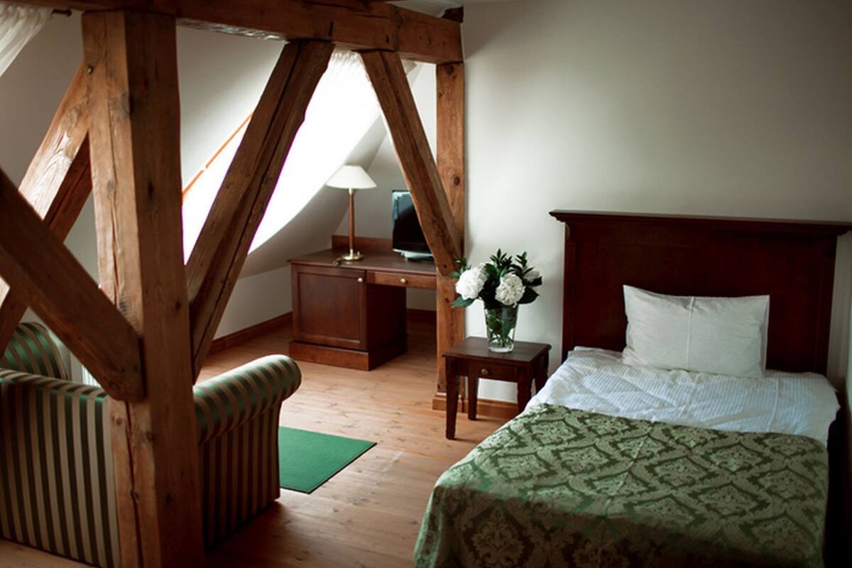 A bedroom with a wooden ceiling, a bed with a patterned bedspread, a nightstand with a lamp, a chair, and a desk with a computer.