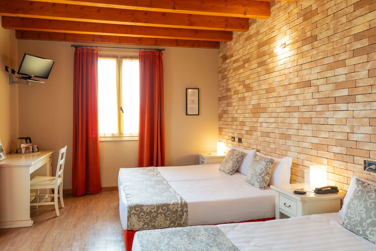 A hotel room with two beds, a desk, a TV, and a brick wall.