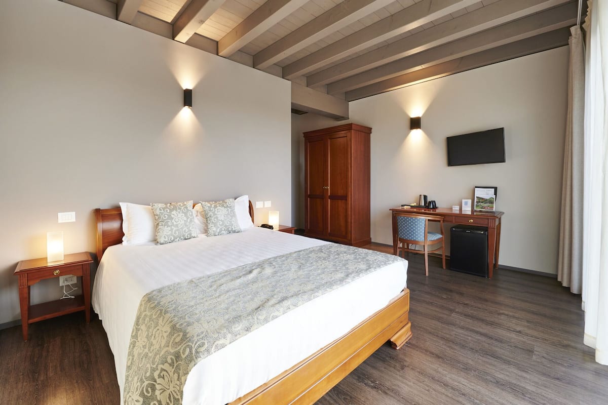 A hotel room with a large bed, wooden headboard, bedside tables with lamps, a wardrobe, a desk with a chair, a television, and a flat-screen TV mounted on the wall.