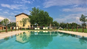 A clear swimming pool with a wooden deck, a stone building, and trees in the background. - Agriturismo La Razza (Reggio nell'Emilia)