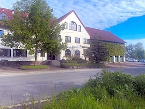 Un bâtiment à deux étages, avec un toit rouge, une enseigne indiquant « Hôtel Spitzer Wirt », et une allée pavée menant à l’entrée.