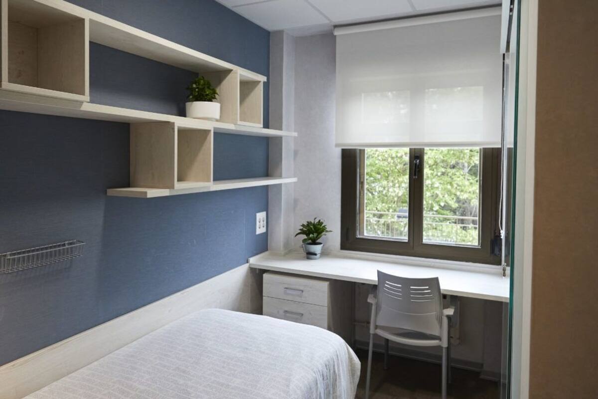 Une chambre à coucher avec un lit, un bureau et une chaise. Il y a des étagères au-dessus du bureau et une fenêtre avec des stores.