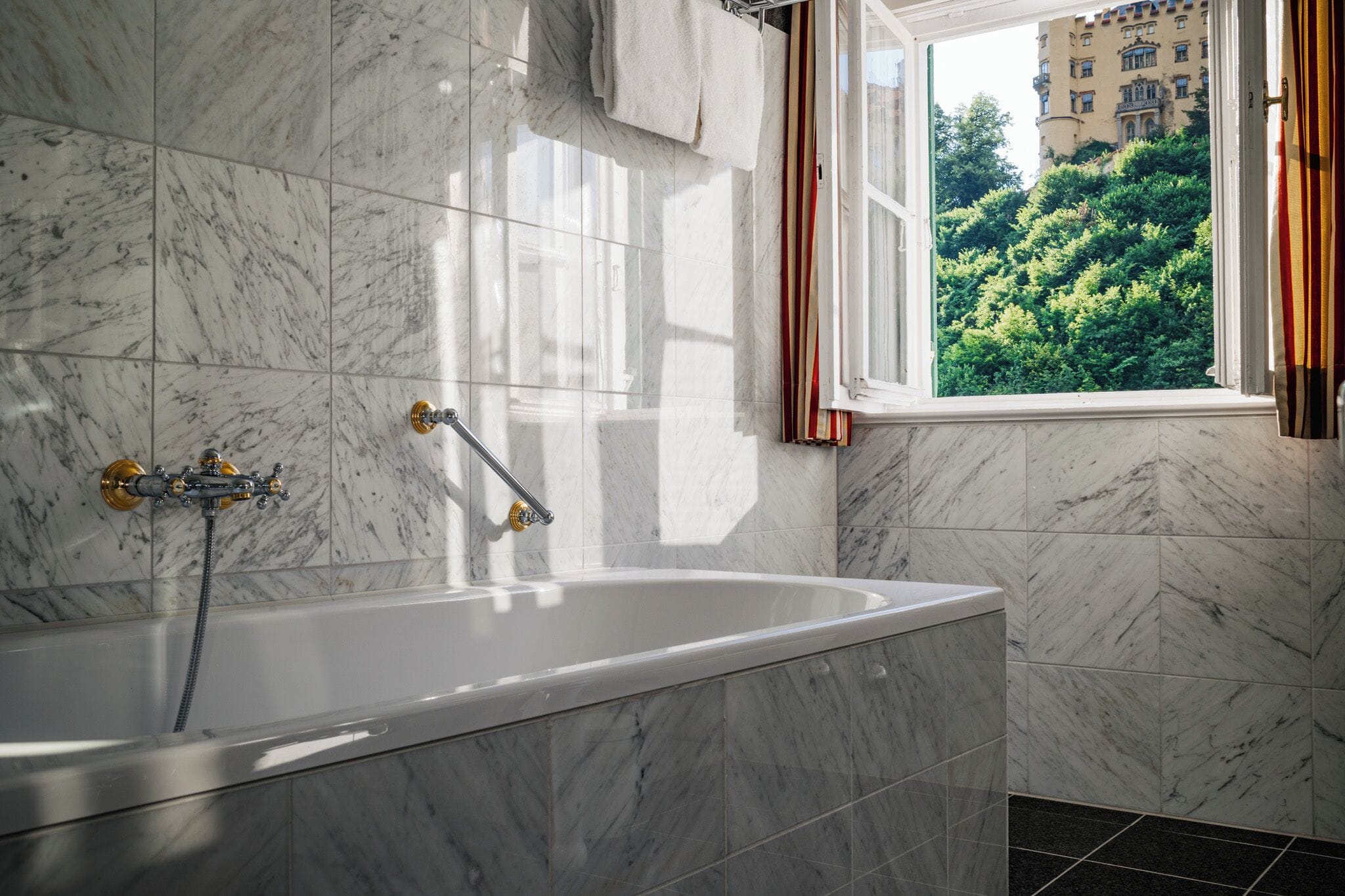 Ein Badezimmer mit Badewanne, Marmorfliesen, einem Fenster mit Vorhängen und Blick auf ein Gebäude und Bäume.
