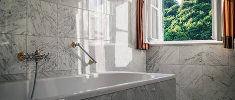 Ein Badezimmer mit Badewanne, Marmorfliesen, einem Fenster mit Vorhängen und Blick auf ein Gebäude und Bäume.