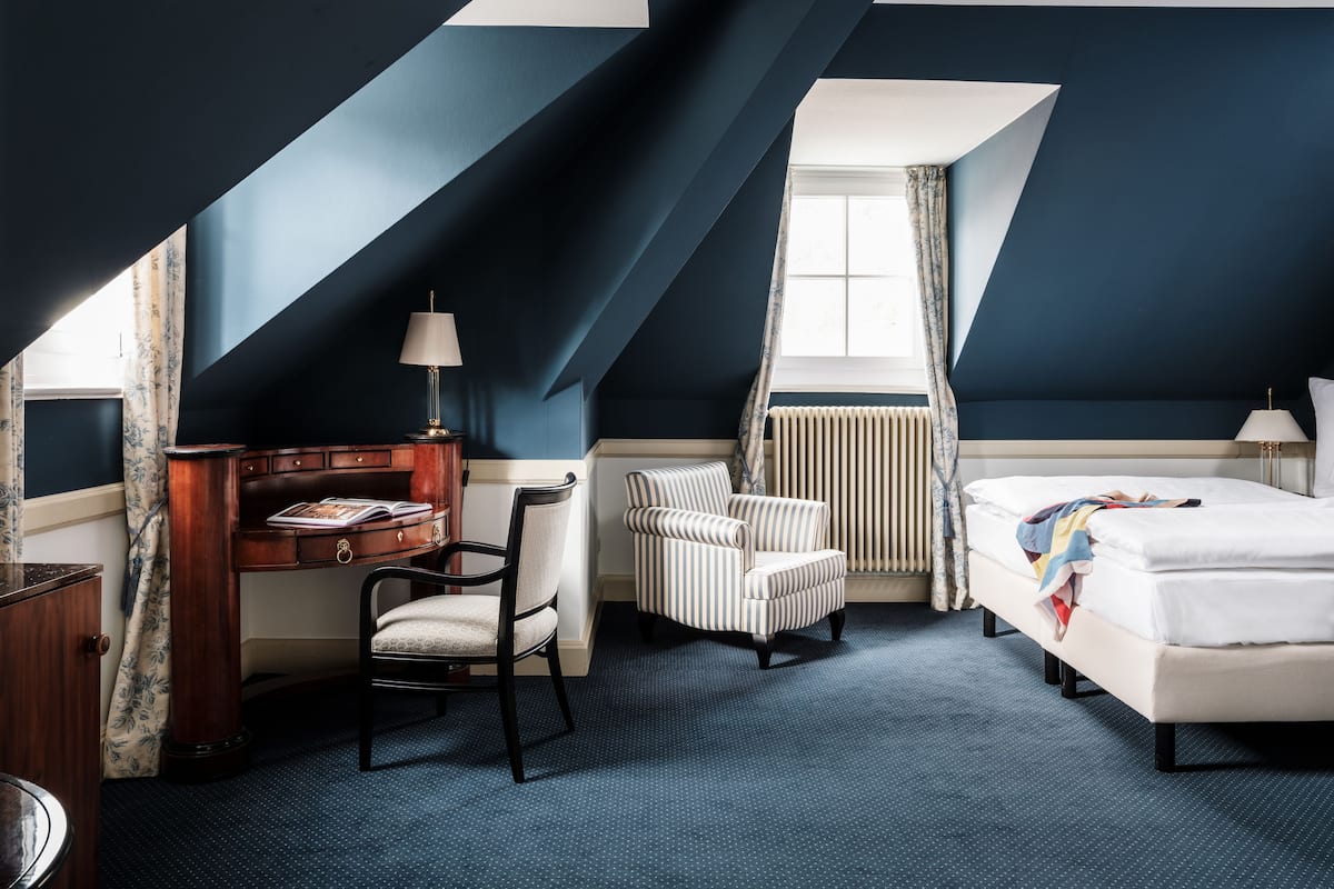 A hotel room with a sloped ceiling, a desk with a lamp, a striped armchair, a bed, and a window with curtains.