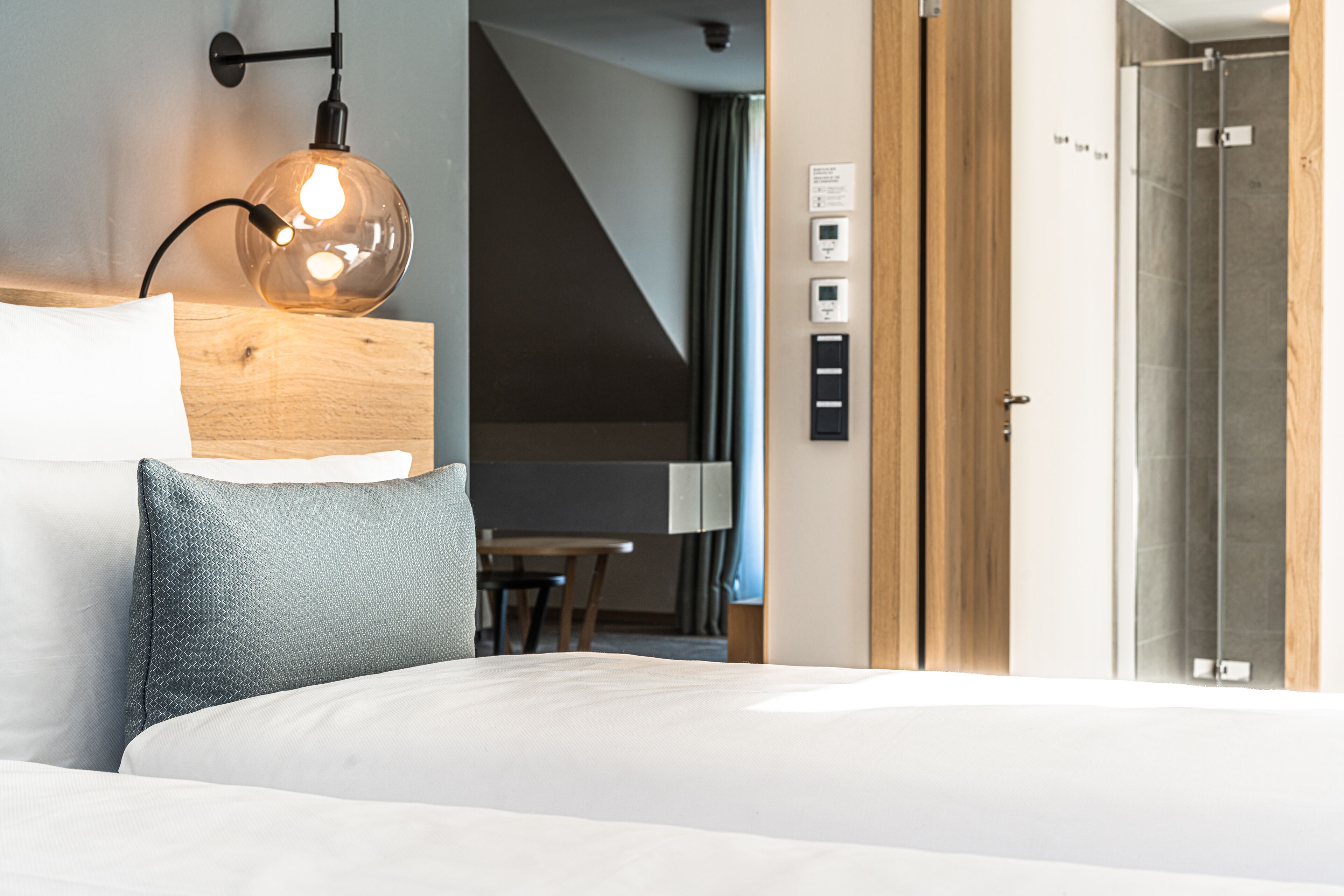 Ein modernes Hotelzimmer mit einem ordentlich bezogenen Bett, einem Holz-Kopfteil, einer Wandlampe und einer sichtbaren Badezimmertür.