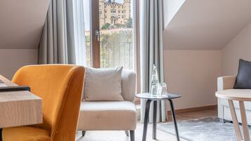 A modern hotel room with a sofa, a small round table with glasses, and a view of a castle through large windows.