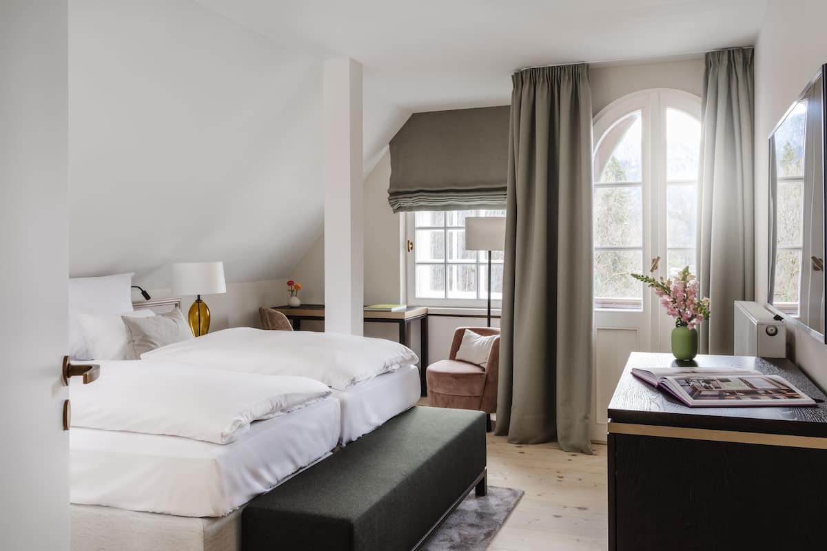 A bedroom with a bed, bedside table, lamp, chair, desk, and a window with curtains.