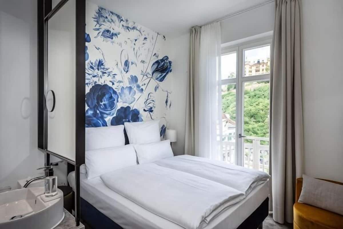A bedroom with a large bed, a bathroom with a sink, and a window with a view of greenery.