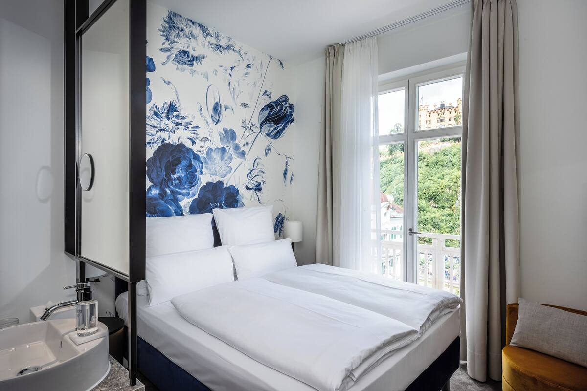 A hotel room with a large bed, a bathroom with a sink, and a window with a view of greenery and buildings.