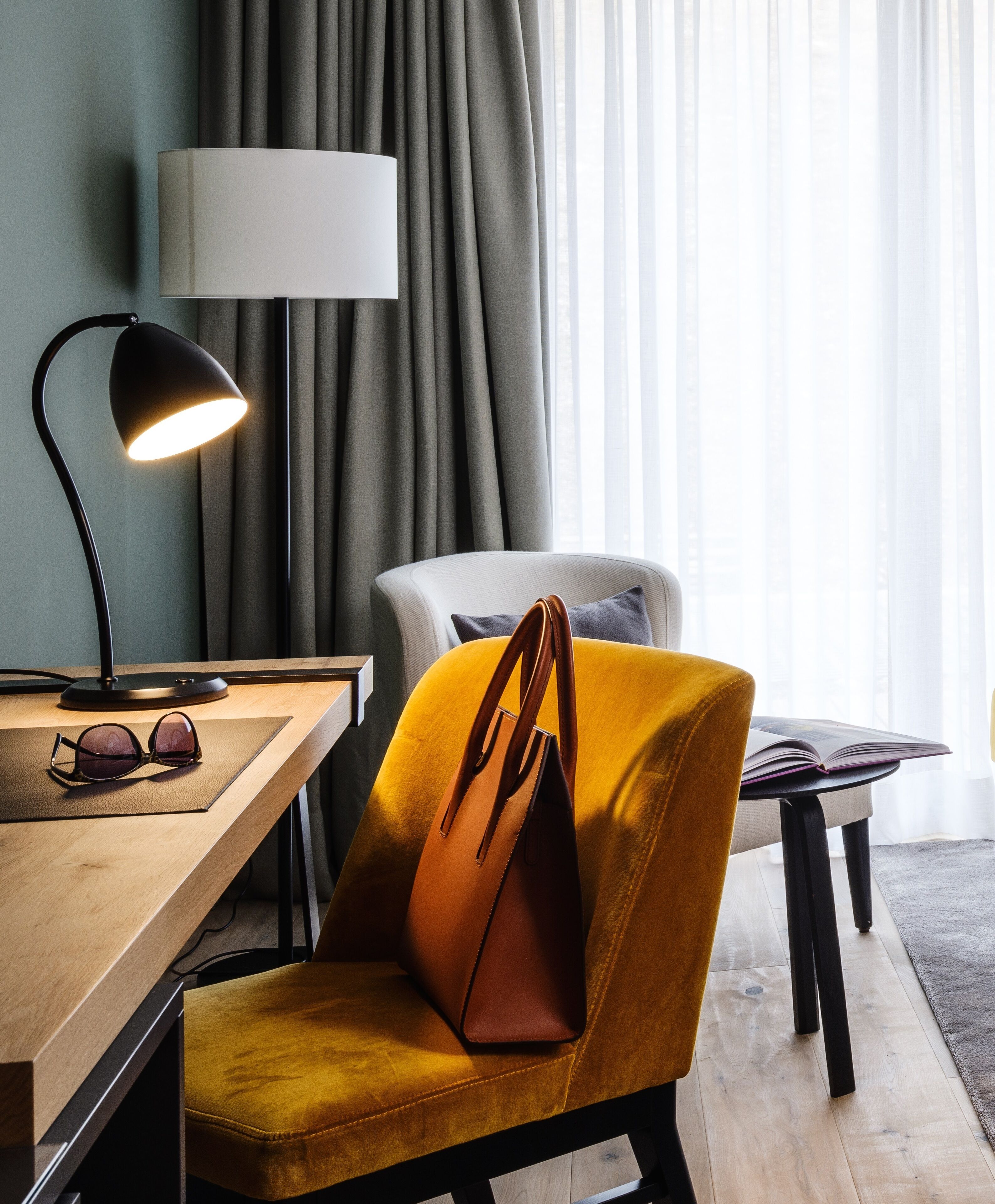 Una habitación moderna con un escritorio de madera, una lámpara, una silla y un bolso marrón, además de un amplio ventanal con cortinas.