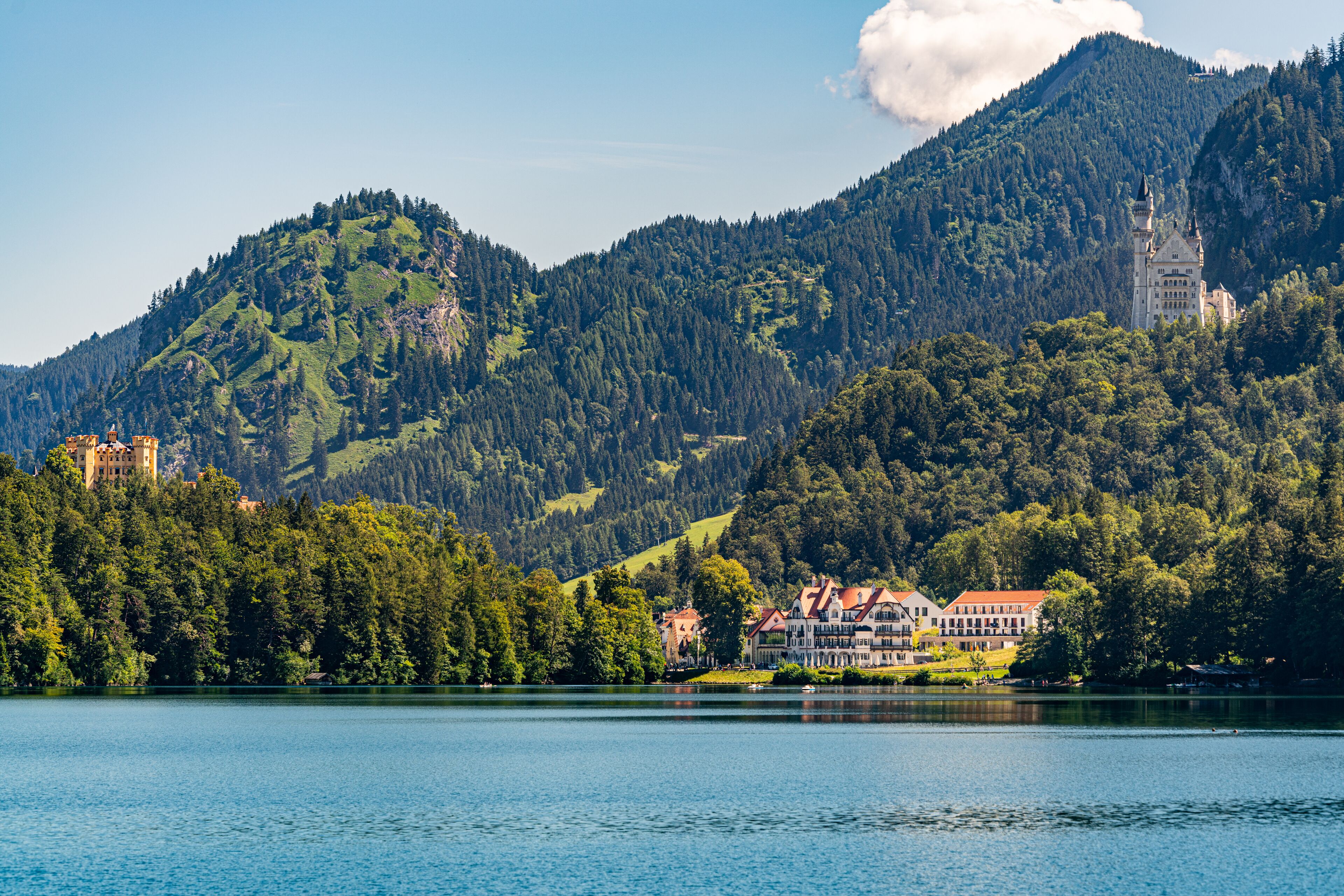 Une vue sur un château au bord d’un lac, entouré de collines verdoyantes et surplombé d’un ciel bleu clair.