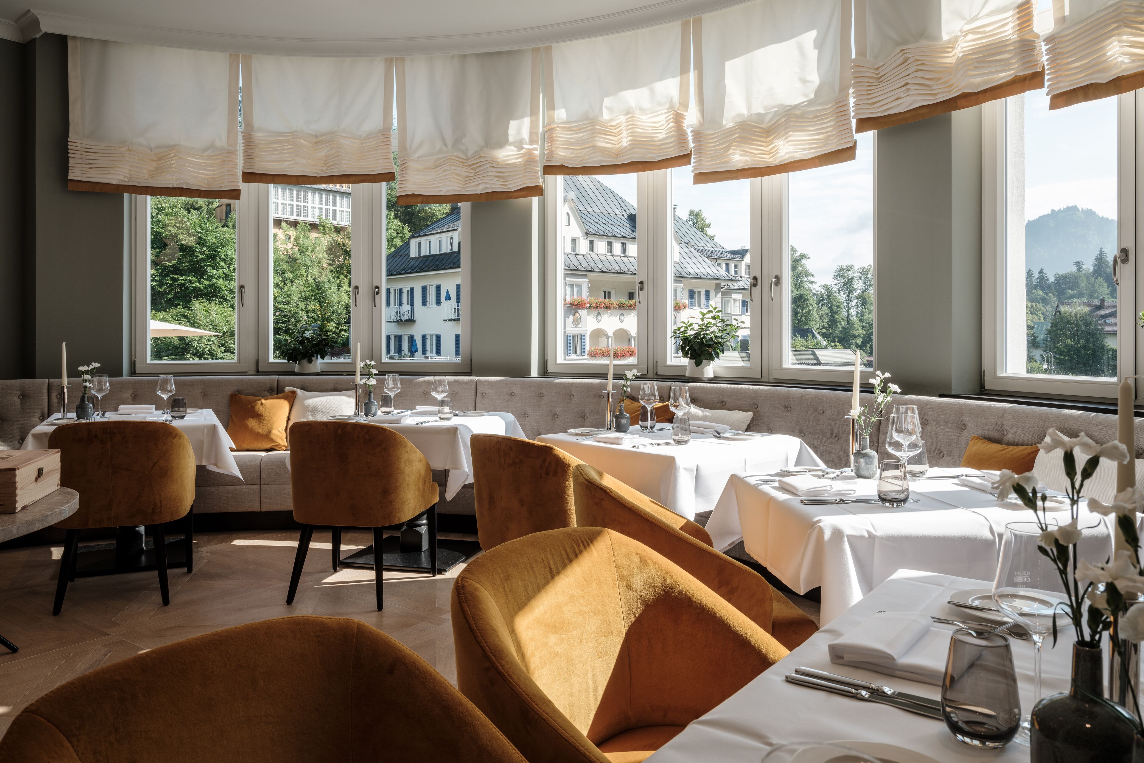 A restaurant with round tables, velvet chairs, and large windows offering a view of buildings and greenery.
