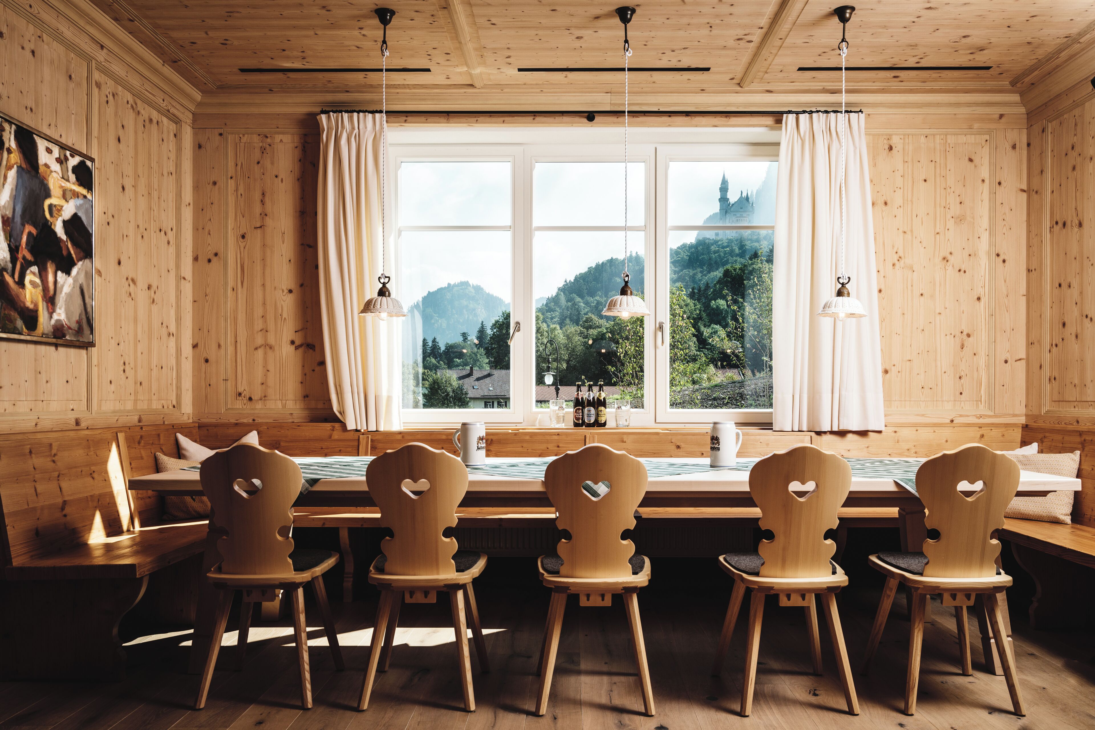 A wooden interior with a dining table and chairs, a window with curtains, and a painting on the wall.