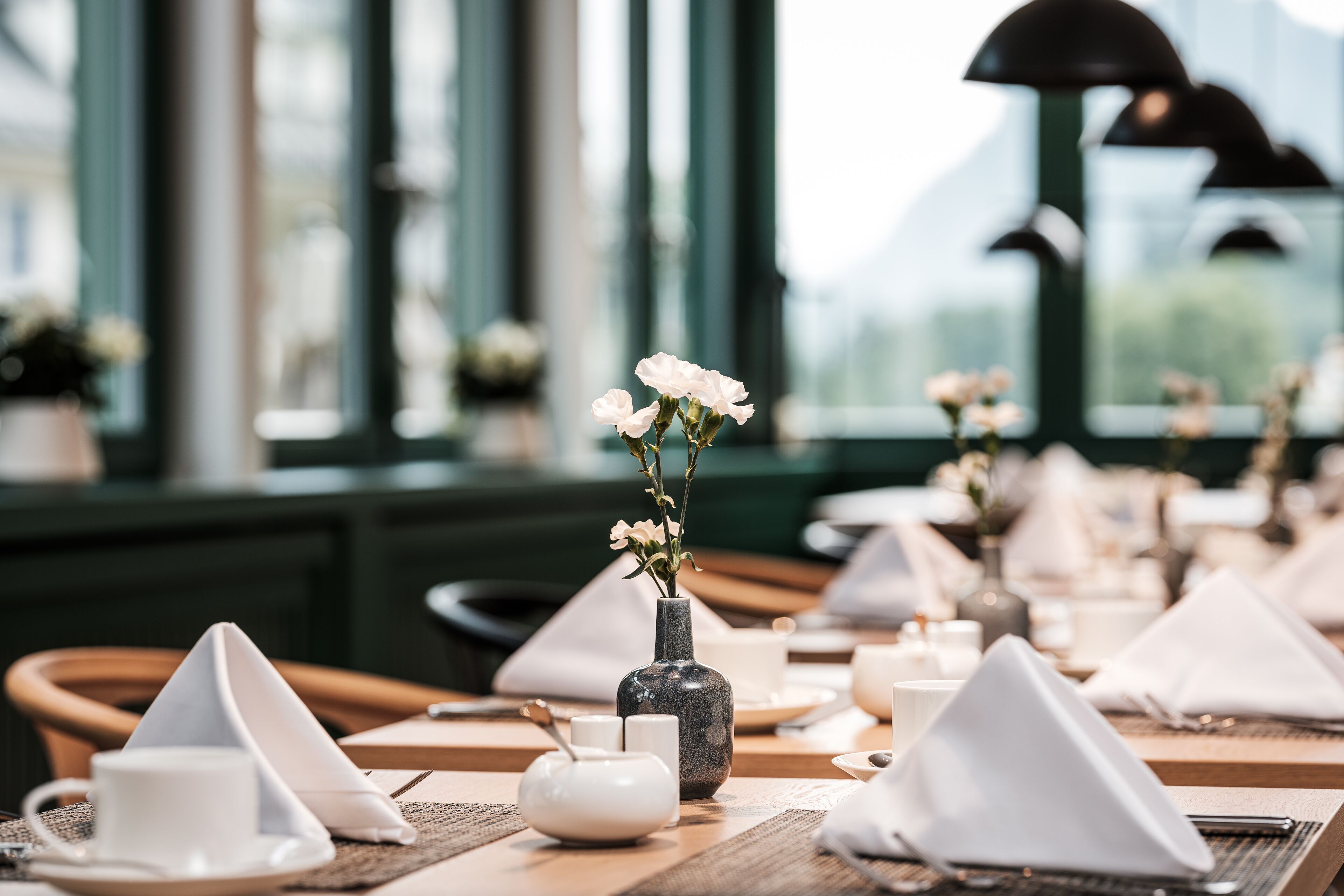 A restaurant setting with neatly arranged tables, each with a white napkin, a small vase with flowers, and a teapot.