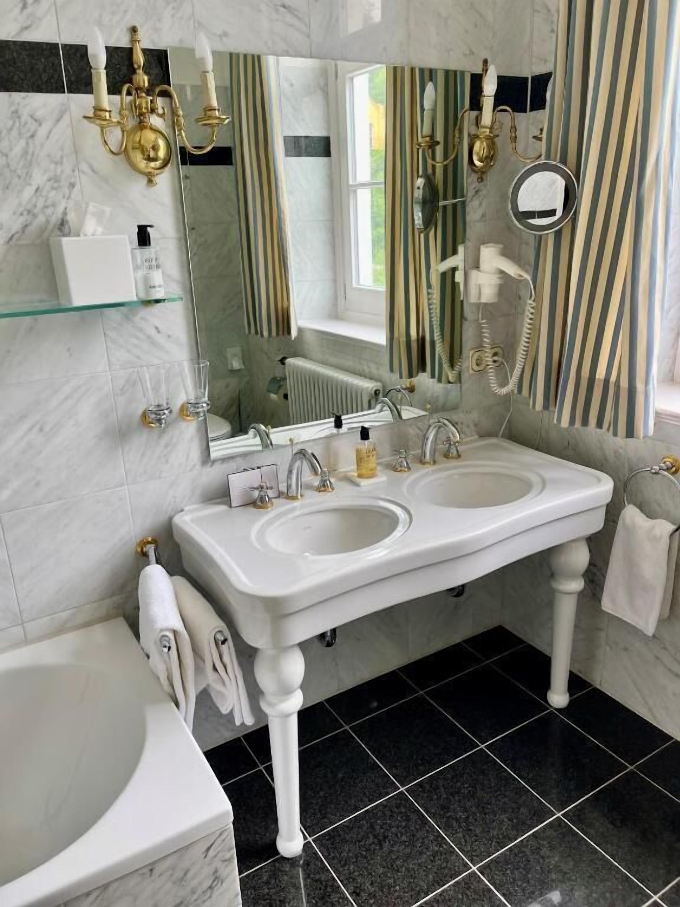 A bathroom with a double sink, a bathtub, a glass shelf with toiletries, a mirror, and a window with striped curtains.